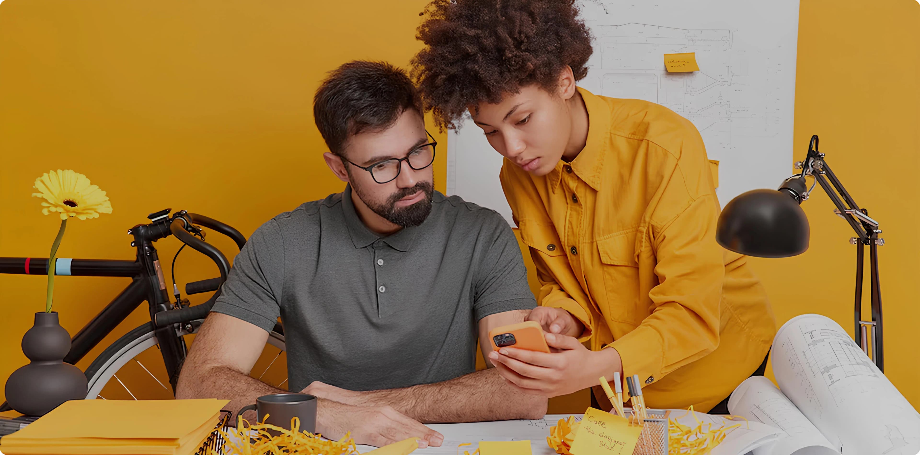 Dois designers ou arquitetos, um homem e uma mulher, examinando algo em um telefone celular em uma sala de escritório com tema amarelo.